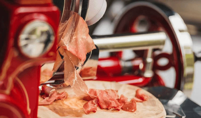 Thinly sliced prosciutto on a wooden board beside a vintage red meat slicer